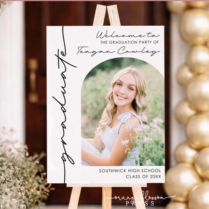 May include: A graduation party welcome sign on a wooden easel. The sign features a photo of a smiling person and the text "Welcome to the Graduation Party of Teagan Cowley." Additional text includes "Southwick High School Class of 20XX."