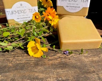 May include: Several bars of turmeric tea soap are displayed with yellow flowers and greenery on a wooden surface. Each bar has a white label with the words "Turmeric Tea" printed on it. The soap has a natural, golden color.