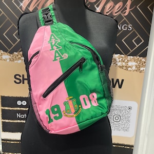May include: A pink and green sling bag with black zippers and mesh side pocket. The bag features the Greek letters AKA and the number 1908 in white. The bag is displayed on a black mannequin.