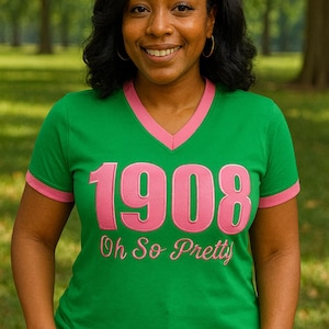 Puede incluir: Camiseta verde con cuello en V y ribete rosa con el texto "1908 Oh So Pretty" en rosa. La camiseta está hecha de un material suave y es adecuada para ropa casual. La imagen está tomada al aire libre.