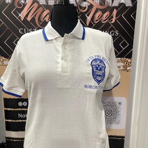 May include: White polo shirt with blue trim on the collar and sleeves. The shirt features the Zeta Phi Beta Sorority crest embroidered on the chest. The shirt has a three-button placket.