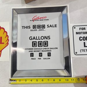 May include: A vintage Gilbarco Calco-Meter gas pump display with a silver frame and white background. The display shows the text "THIS SALE", "DOLLARS CENTS", "GALLONS", and "CUSTOMER SHOULD CHECK GALLON AND MONEY READINGS". There are three digital displays showing "000" for the sale amount, gallons, and price per gallon.