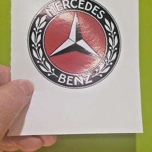 May include: A white sticker featuring the Mercedes-Benz logo. The logo is a black and white three-pointed star inside a red circle, surrounded by a black ring with the words "MERCEDES" and "BENZ". The sticker is held against a green background.
