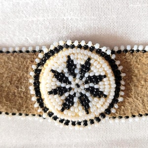May include: A tan suede choker necklace with a central circular beaded design. The design features a white background with a black starburst pattern. The edges are trimmed with white and black beads.