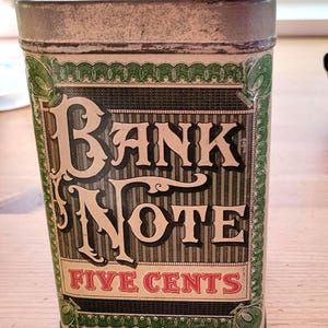 May include: Vintage metal tin with a rectangular shape and a hinged lid. The tin features the words "BANK NOTE" in large, ornate letters, with "FIVE CENTS" below. The design includes green and black accents.