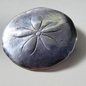 May include: A silver-toned metal pendant shaped like a sand dollar with a detailed starburst design.