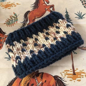 May include: A chunky knit headband in navy blue, cream, and tan. The headband features a geometric pattern and is made of thick yarn. The background is a patterned fabric with horses and desert plants.