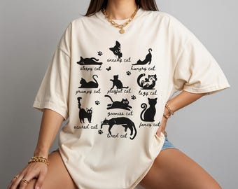 May include: Cream-colored t-shirt featuring black cat illustrations with various poses and labels like "sleepy cat" and "hungry cat." The shirt is worn by a person. The shirt has a relaxed fit.