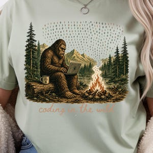 May include: Sage green t-shirt with a Bigfoot illustration coding on a laptop in a forest scene. A binary code stream floats above a campfire. The text "coding in the wild" is below the illustration.