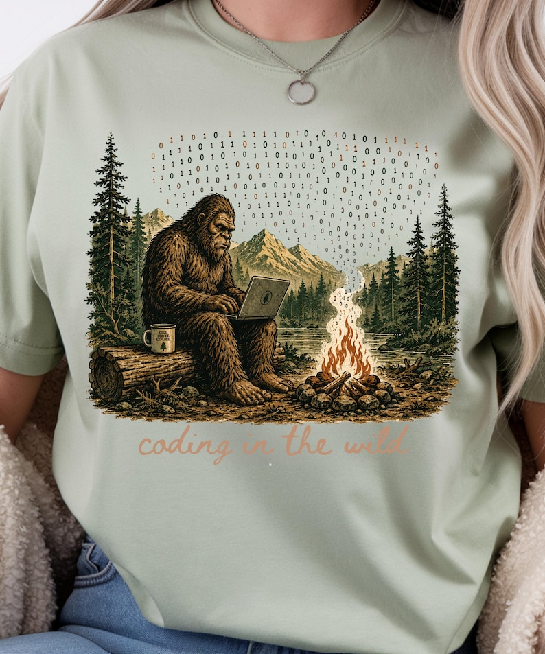 May include: Sage green t-shirt with a Bigfoot illustration coding on a laptop in a forest scene. A binary code stream floats above a campfire. The text "coding in the wild" is below the illustration.