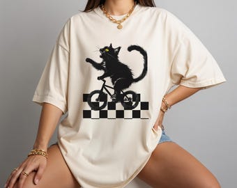 May include: Cream-colored t-shirt featuring a black cat riding a bicycle. The cat is depicted with yellow eyes and an open mouth, set against a black and white checkered pattern. The shirt has short sleeves and a relaxed fit.