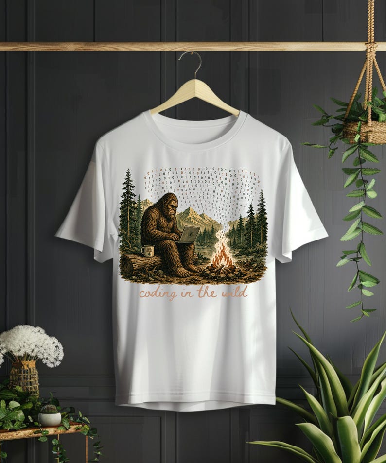 May include: White t-shirt with a Bigfoot graphic using a laptop in a forest scene. The design includes a campfire, mountains, trees, and the text "coding in the wild".
