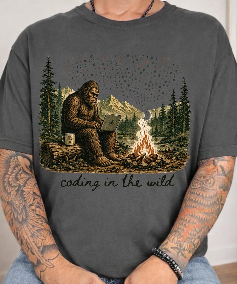 May include: Dark gray t-shirt featuring a Bigfoot character coding on a laptop in a forest setting. The design includes a campfire, mountains, trees, and the text "coding in the wild".