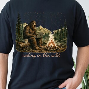 May include: Navy blue t-shirt with a Bigfoot illustration coding on a laptop in a forest. The design includes a campfire, mountains, trees, and the text "coding in the wild."