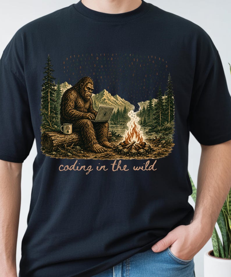 May include: Navy blue t-shirt with a Bigfoot illustration coding on a laptop in a forest. The design includes a campfire, mountains, trees, and the text "coding in the wild."