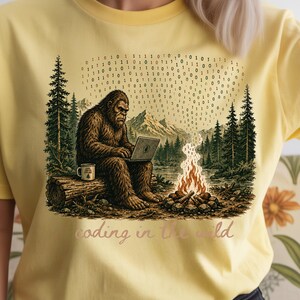 May include: Yellow t-shirt with a Bigfoot graphic coding on a laptop in a forest scene. The design includes a campfire, mountains, and binary code. The text "coding in the wild" is also present.