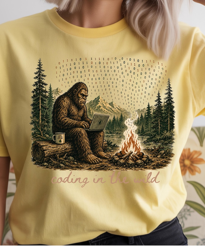 May include: Yellow t-shirt with a Bigfoot graphic coding on a laptop in a forest scene. The design includes a campfire, mountains, and binary code. The text "coding in the wild" is also present.