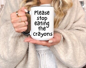 May include: White ceramic mug with black handle and rim, featuring the text "Please stop eating the crayons" in black. The mug is held by a person wearing a beige sweater and gold rings.
