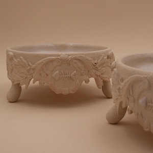 May include: Two round, beige ceramic bowls with a textured, matte finish. Each bowl features ornate, raised scrollwork detailing along the exterior rim and stands on three short, rounded feet. A small fish skeleton relief is centred on the front.