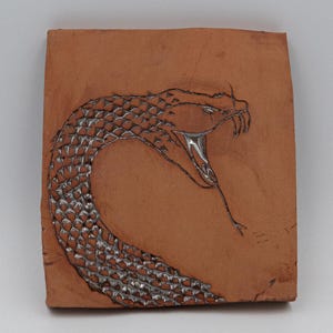 May include: A square, terracotta-coloured tile featuring a detailed, open-mouthed snake design. The snake's scales are rendered in a silver, textured pattern, with a silver tongue extending from its mouth. The artwork is centered on the tile.