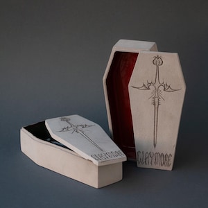 Puede incluir: Dos cajas de cerámica de color blanco roto con forma de ataúd y sus tapas. Cada tapa presenta un diseño de espada grabado y la palabra CLAYMORE. Una caja está de pie, revelando un interior rojo brillante, mientras la otra está tumbada.