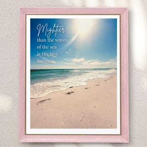 Könnte beinhalten: Ein gerahmter Druck mit einer Strandszene mit strahlend blauem Himmel und Ozean. Der Druck enthält den Text "Mightier than the waves of the sea is His love for you" in eleganter Schrift. Der Rahmen ist zartrosa.