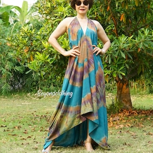 Op de afbeelding: Een vrouw draagt een tealblauwe en bruine tie-dye maxi-jurk met een halternek. De jurk heeft een high-low zoom en is gemaakt van een zachte, vloeiende stof. De vrouw staat op een grasveld met bomen op de achtergrond.