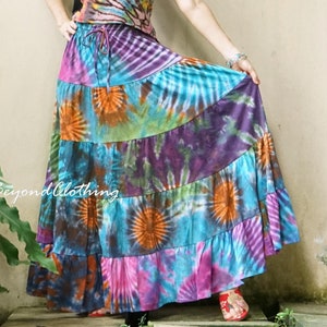 May include: A woman wearing a colorful tie-dye tank top and a long, multi-colored tie-dye skirt. The skirt has a layered, ruffled design. The woman is standing in front of a white wall with green plants in the background. The text "Beyond Clothing" is visible in the lower left corner of the image.