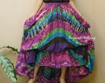 May include: A woman wearing a black t-shirt with a small design on the front and a long, multi-colored tie-dye skirt. The skirt has a layered design and is flowing. The woman is standing in front of a beige wall with green plants in the background.