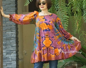 May include: A woman wearing a long-sleeved, purple, orange, and blue tie-dye dress with a ruffled hem. She is wearing blue jeans and brown sandals. The dress has a square neckline and is loose-fitting.