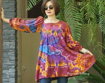 May include: A woman wearing a tie-dye tunic with long sleeves and a gathered skirt. She is wearing blue jeans and sandals. The tunic is a vibrant mix of orange, purple, and blue.