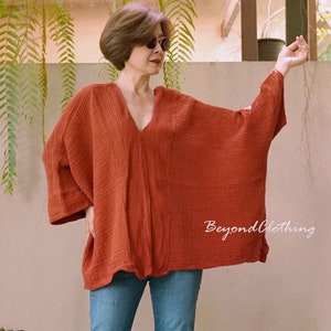 May include: Rust-colored, loose-fitting top with a deep V-neck and wide sleeves. The top is made of a textured fabric and is paired with blue jeans. The text "Beyond Clothing" is visible on the bottom right.