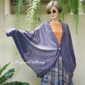 May include: A purple, oversized, long-sleeved cardigan with a single button closure, worn over an orange top and a blue and brown tie-dye patterned skirt. The text BeyondClothing is printed in white on the front of the purple cardigan.