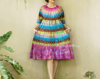 May include: A woman wearing a colorful tie-dye dress with a ruffled hem. The dress has a rainbow pattern with shades of pink, blue, yellow, and brown. She is wearing sunglasses and red sandals.