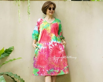 May include: A woman wearing a tie-dye dress with a V-neck and 3/4 sleeves. The dress has a red, green, yellow, and white color scheme. The woman is standing in front of a white wall with green plants on either side.