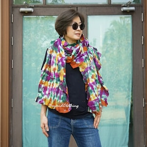 Può includere: Una donna indossa una camicia nera e jeans blu con una sciarpa colorata tie-dye intorno al collo. La sciarpa ha sfumature di viola, verde, arancione e bianco.