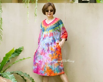 May include: A woman wearing a colorful tie-dye dress with a V-neck and short sleeves. She is standing in front of a white wall with green plants hanging down. The dress has a large pocket on each side. The woman is wearing red sandals and sunglasses.