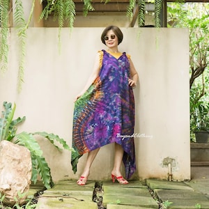 May include: A woman wearing a purple, green, and orange tie-dye dress with a high-low hemline. The dress has a V-neck and is sleeveless. She is wearing sunglasses and red sandals. The dress is made by Beyond Clothing.