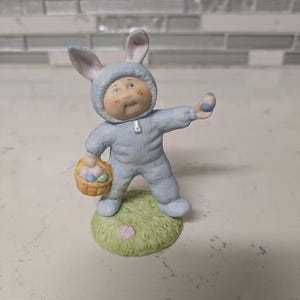 May include: A porcelain figurine of a Cabbage Patch Kid dressed as a bunny holding a basket of Easter eggs. The figurine is wearing a blue bunny costume with a white lining and has a basket of colorful eggs in one hand and a single blue and yellow egg in the other.
