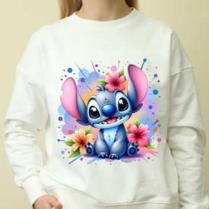 Puede incluir: Sudadera blanca con un personaje de dibujos animados azul y rosa con ojos grandes y una corona de flores. El personaje está rodeado de salpicaduras de pintura de colores y flores.