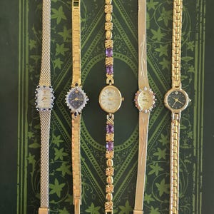 May include: Five gold-toned wristwatches with various band styles and face designs are displayed. The watches feature rectangular, oval, and round faces, some adorned with gemstones. The bands include mesh, link, and patterned styles. The watches are numbered 1 through 5.