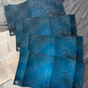 May include: Folded blue paper with a starry night design. The paper has a gradient effect, transitioning from dark blue to lighter shades. White dots represent stars. The corner of the paper has a small logo. Beside the paper are old book pages.