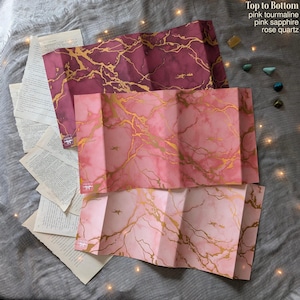 May include: Three folded paper maps with a marble design in shades of pink and gold. The top map is a deep pink, the middle is a lighter pink, and the bottom is a pale pink. The maps are folded and have a textured appearance. The text "Top to Bottom pink tourmaline pink sapphire rose quartz" is visible.