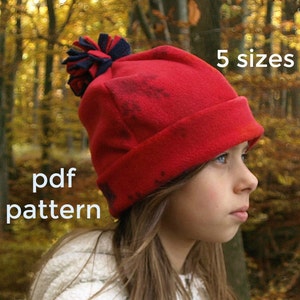 May include: A red fleece hat with a blue and white pom pom. The hat is available in 5 sizes and comes with a PDF pattern. The hat is being worn by a young person with long brown hair.
