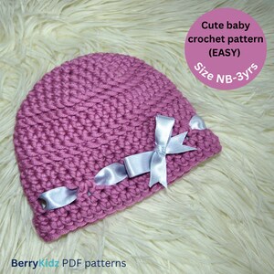 May include: A pink crocheted baby hat with a white satin bow. The hat is on a white fluffy surface. The text "Cute baby crochet pattern (EASY) Size NB-3yrs" is in the upper right corner of the image.