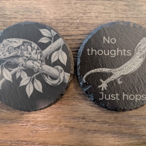 May include: Two round, dark gray slate coasters. One coaster features a detailed engraving of a gecko on a branch with leaves. The other coaster has the text "No thoughts Just hops" with a gecko illustration.