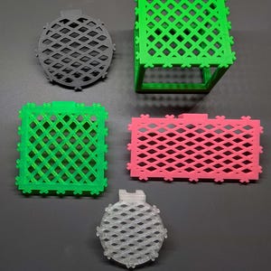 May include: An assortment of 3D-printed geometric shapes in various colors. The collection includes a gray circular ornament, a green square table, a green square, a pink rectangle, and a clear circular ornament, all with a lattice design.