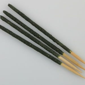 May include: Five dark green incense sticks with light brown tips are arranged diagonally against a white background. The incense sticks have a textured appearance and are likely scented. The tips are a natural wood color.