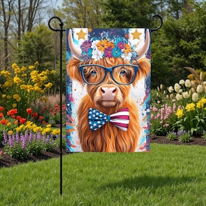 May include: A decorative garden flag featuring a whimsical illustration of a Highland cow. The cow wears blue glasses, a floral crown, and a patriotic bow tie. The flag is displayed on a black metal stand in a garden setting.