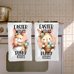 Puede incluir: Dos paños de cocina blancos con textura de gofre que presentan un gráfico de un conejo de dibujos animados sosteniendo un huevo de Pascua de colores. El texto "EASTER WISHES" aparece sobre el conejo y "BUNNY KISSES" debajo, con flores y una cesta.